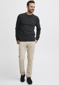 Pulover tricotat din cărbune cu guler rotund, pantaloni bej slim-fit și pantofi negri, având un mic detaliu cu logo pe pulover.