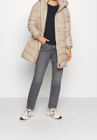 Beige pufferjas met een met bont gevoerde capuchon, drukknoopsluiting en geribbelde manchetten, gecombineerd met grijze spijkerbroek en witte sneakers.