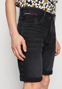 Pantalones cortos de denim negros con dobladillos enrollados, que presentan un diseño clásico de cinco bolsillos y costuras de contraste. Mano visible colocada sobre el muslo.