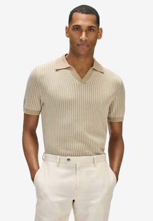 Mann trägt ein beige-farbenes kurzärmeliges Strick-Poloshirt mit vertikalen Streifen, in hellbeige Hose gesteckt, Hände in den Taschen, neutraler Hintergrund.