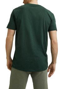 Homme portant un t-shirt vert foncé à manches courtes et un pantalon vert olive, vu de dos sur un fond blanc.