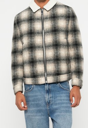 Homme portant une veste zippée à motif à carreaux noir, gris et beige avec un col sherpa blanc, associée à un jean denim bleu clair.