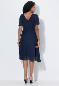 Marineblaues Kleid mit durchsichtigen Schmetterlingsärmeln, V-Ausschnitt und einem ausgestellten Rock, aus leichtem Stoff gefertigt. Kombiniert mit silbernen Absatzsandalen.