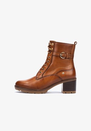 Botte de cheville en cuir marron avec un bout rond, un laçage sur le devant, des garnitures en ton or et un talon épais. Présente un détail de boucle sur le côté.