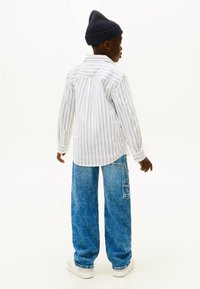 Striped white and blue button-up shirt, loose blue denim jeans, and a dark knit beanie. White sneakers complete the outfit.