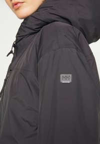 Chaqueta impermeable negra con un alto cuello, capucha con cordón y parche con logo en la manga. Textura suave, diseño moderno y forma ajustada.