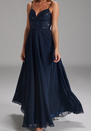 Kobieta w granatowej wieczorowej sukni bez rękawów z cekinowym gorsetem i lejącym się tiulowym dołem, stojąca na tle jednolitego szarego tła.