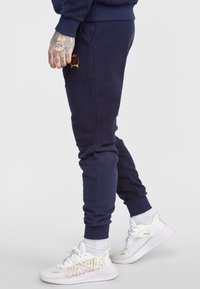 Námořnicky modré tepláky z měkkého materiálu s žebrovanými manžetami, bočními kapsami a malým vyšitým logem na přední straně. Bílé tenisky s logem.