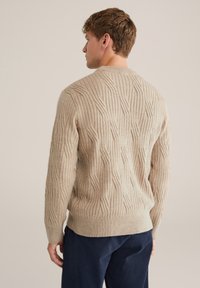 Beige kötött pulóver bordázott textúrával és geometriai mintázattal, kerek nyakkivágással és szűkített mandzsettákkal, sötét nadrág felett viselve.