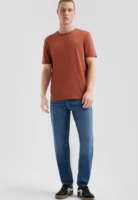 Roodbruine t-shirt met korte mouwen en een ronde halslijn, gecombineerd met blauwe rechte jeans en zwarte sneakers met witte accenten.