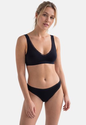 Giovane donna in piedi, che indossa un reggiseno nero con rifiniture in pizzo e mutandine abbinate con rifiniture in pizzo nero, su uno sfondo neutro.