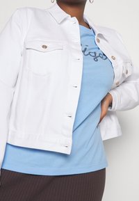 Weiße Jeansjacke mit goldenen Knöpfen und zwei Brusttaschen, getragen über einem hellblauen T-Shirt mit schwarzer Stickerei.