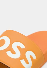 Oransje slidesandal med bred stropp med fet hvit tekst. Jevn tekstur og formet fotseng for komfort.