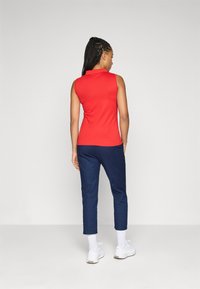 Donna con capelli intrecciati che indossa un top rosso senza maniche, pantaloni blu navy, calzini bianchi e sneakers bianche, in piedi su uno sfondo chiaro e semplice.