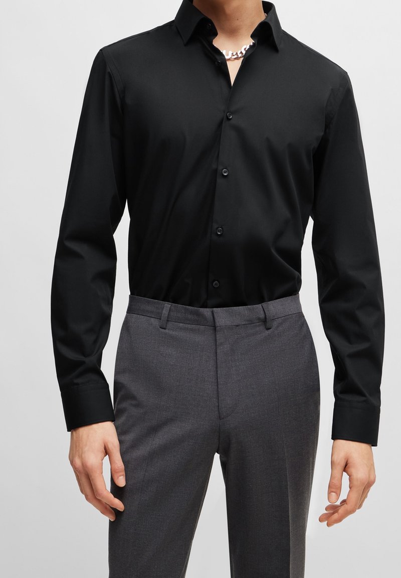 Homme portant une chemise noire à manches longues boutonnée, rentrée dans un pantalon de costume gris, avec une chaîne en argent visible au col.