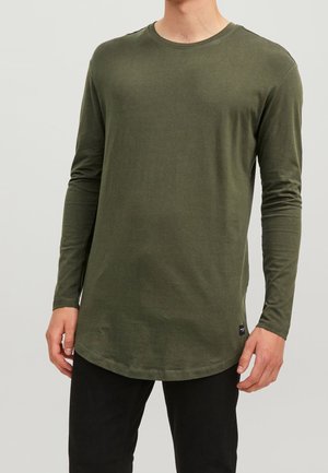 Man som bär en olivgrön långärmad tröja med rund hals och svarta byxor mot en neutral bakgrund.