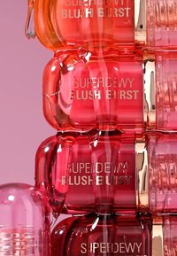 Opgestapelde cosmeticacon containers gemarkeerd met "Superdewy Blush Burst." Ze hebben glanzende plastic, ronde vormen, in tinten van rood en roze met gouden accenten.