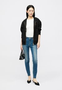 Mulher a usar um hoodie preto com fecho, blusa branca, jeans skinny azuis, sapatos pretos de tacão pequeno, segurando uma pequena mala preta, em pé contra um fundo branco.