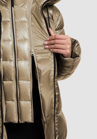 Opvouwbare jas in reflecterend lichtgoud materiaal. Voorzien van een capuchon, een doorgestikt ontwerp en dubbele ritsen met zwarte accenten. Er is een textuur zichtbaar op het oppervlak.