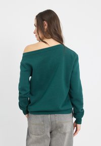 Sudadera verde sin hombros con mangas largas, puños acanalados y un corte relajado, combinada con jeans sueltos de color gris claro.