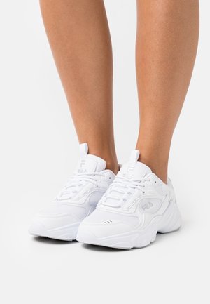 Gros plan sur des jambes portant des baskets blanches FILA avec des semelles épaisses, des lacets et des détails en mesh sur un fond blanc uni.