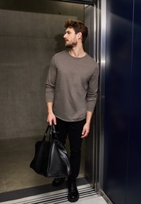 Grå långärmad shirt, svarta byxor och svart läderväska. Står i en hiss med metallaccenter och betongväggar.