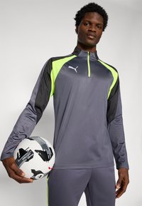 Completo sportivo grigio e nero con vivaci accenti in verde neon, dotato di colletto a mezza zip. Il modello tiene in mano un pallone da calcio a motivi in bianco e nero.