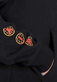 Felpa con cappuccio nera in tessuto morbido con polsini a coste. Presenta disegni di globi gialli e scritte rosse lungo la manica.