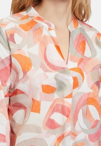 Lyhythihainen, kevyt paita, joka on valmistettu kankasta, jossa on akvarellipainatus, jossa on abstrakteja oransseja, vaaleanpunaisia ja harmaita muotoja valkoisella taustalla.