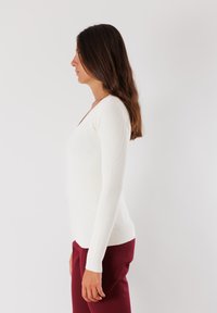 Maglietta bianca a maniche lunghe a coste con scollo a V, silhouette aderente e texture liscia. Abbinata a pantaloni borgogna.