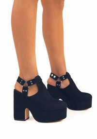 Scarpe platform in suede nero con tacco alto a blocco e una cinghia con dettagli in argento. Design aperto sul retro per una facile indossabilità.
