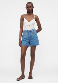 Femme aux cheveux tressés portant un top blanc sans manches et un short en jean bleu avec broderies florales, debout devant un fond uni.