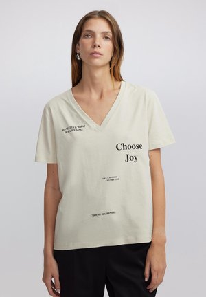 Helle beigefarbene Baumwoll-T-Shirt mit V-Ausschnitt, das verschiedene Textdrucke in Schwarz zeigt, darunter "Wähle Freude" und "Egal, was passiert".