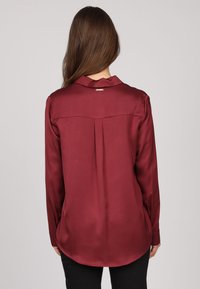 Blouse bordeaux à manches longues avec un col, texture lisse, détail de plis au dos et accent de marque subtil juste en dessous du col.