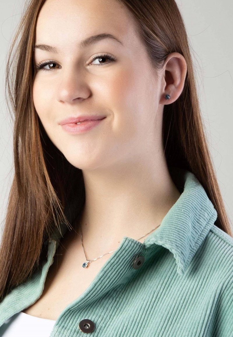 Lichtgroene corduroy jas met een omgeslagen kraag, voorzien van donkere knopen; zilveren ketting met een blauw edelsteen hanger zichtbaar.