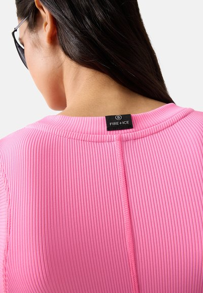 Kvinde med langt mørkt hår iført lyserød ribstrikket skjorte med sort Fire+Ice-mærke på bagsiden af halsudskæringen, vist bagfra
