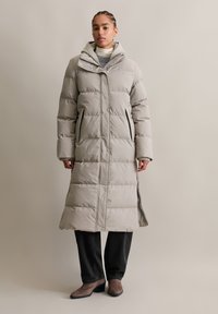 Lange pufferjas in lichtgrijs met een gewatteerd ontwerp, zijzakken en een capuchon. De jas reikt tot halverwege het scheenbeen voor extra warmte.
