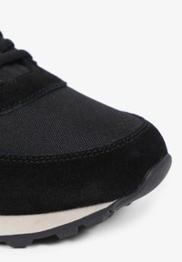 Zwarte sportieve schoen met een combinatie van suede en mesh materialen, gevormde rubberen loopzool en een getextureerd oppervlak voor grip.