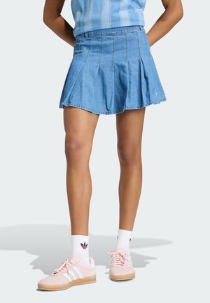 Femme portant une mini-jupe plissée en denim, un haut bleu clair, des chaussettes blanches avec un logo bordeaux, et des baskets roses avec des rayures blanches.