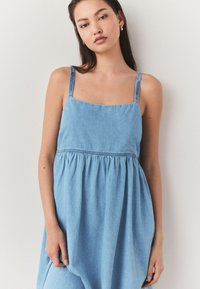 Tânără cu păr lung și închis la culoare, purtând o rochie fără mâneci din denim albastru deschis, pe un fundal simplu, deschis la culoare.