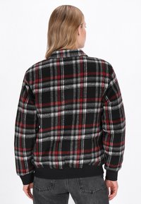 Veste à carreaux noire, rouge et blanche avec un col, des poignets côtelés et une ceinture, fabriquée dans un tissu doux et texturé avec une coupe décontractée.