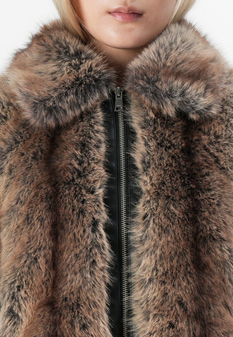 Zbliżenie osoby w brązowo-czarnym futrze sztucznym z dużym kołnierzem i widocznym srebrnym zamkiem błyskawicznym na czarnej odzieży pod spodem.