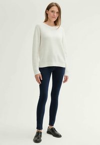 Fehér kötött pulóver kerek nyakkivágással, sötétkék szűk farmerrel és fekete fűzős cipővel párosítva. Egyszerű kivitel, látható minták nélkül.