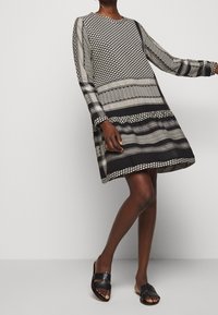 Schwarz-weiß gemustertes Kleid mit langen Ärmeln, rundem Ausschnitt und ausgestelltem Saum. Kombiniert mit schwarzen flachen Sandalen.