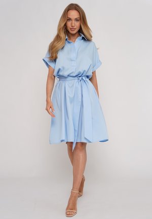 Kobieta idąca do przodu, ubrana w jasnoniebieską sukienkę koszulową z krótkim rękawem, z wiązanym w talii paskiem i beżowymi, otwartymi na palce szpilkami na jednolitym tle.