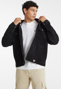 Giubbotto in denim nero con chiusura a bottoni, tasche sul petto e dettaglio del logo triangolare rosso. Indossato sopra una maglietta bianca e pantaloni beige.