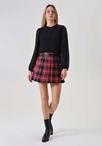Schwarze Bluse mit durchsichtigen Ä RMELN, kombiniert mit einem roten karierten Faltenrock, der einen schwarzen Gürtel hat. Das Model trägt knielange schwarze Stiefel.