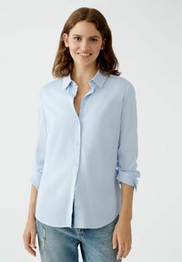 Light blue button-up shirt with long sleeves, soft fabric, chest pocket, and a rounded hem, paired with distressed blue jeans.