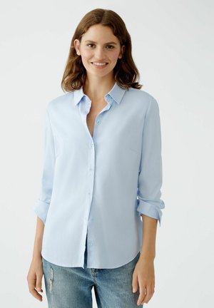 Light blue button-up shirt with long sleeves, soft fabric, chest pocket, and a rounded hem, paired with distressed blue jeans.