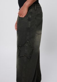 Dark green denim pants with a star-shaped patch and frayed edges. Features a relaxed fit and side pockets. Classic five-pocket design.